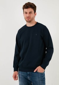 Námornícky modrý sveter z mäkkej tkaniny, s okrúhlym výstrihom a rebrovanými manžetami. Minimalistický dizajn s malým okrúhlym logom na hrudi.