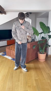 Chaqueta de cremallera gris claro con un patrón de tie-dye, combinada con pantalones de pana oscuros. Zapatillas plateadas y plantas de interior en el fondo.