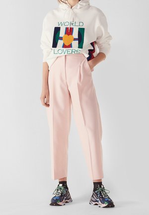 Osoba ubrana w białą bluzę z kapturem z napisem "World Lovers", różowe spodnie z wysokim stanem oraz wielokolorowe masywne trampki z zielonymi sznurówkami.