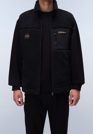 Personne portant une veste polaire noire avec des panneaux avant texturés, une poche poitrine zippée et des écussons de marque ronds et rectangulaires.