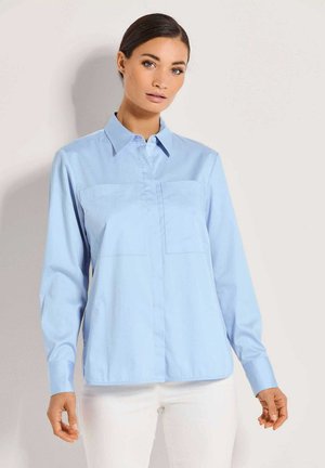 Een vrouw in een lichtblauwe button-up shirt met lange mouwen en een spitse kraag, gecombineerd met witte broek tegen een effen achtergrond.