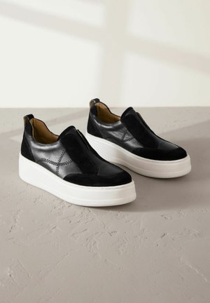 Juodos zomšos ir odos slip-on bateliai su balta platforma, dekoratyvinėmis siuvimo detalėmis ir apvalia nosimi.