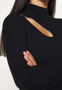 Femme portant un haut noir à manches longues côtelé avec un col montant et une découpe diagonale au niveau de la poitrine, les bras croisés.