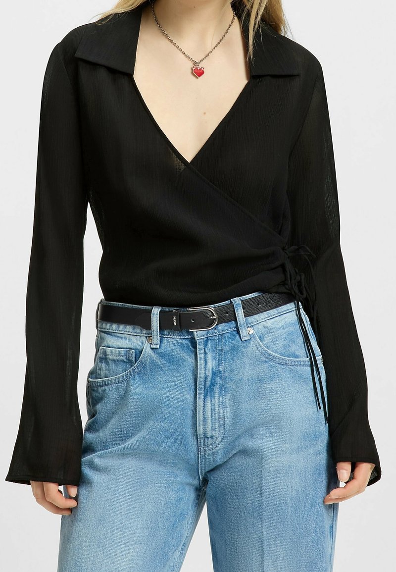 Blouse noire portefeuille à manches longues et col en V profond, associée à un jean taille haute bleu clair et une ceinture noire.