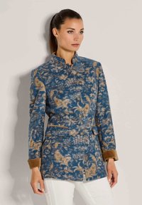 Vrouw in een blauw en tan patroon jasje met knopen en bruine manchetten, gecombineerd met een witte broek, staande tegen een beige muur.