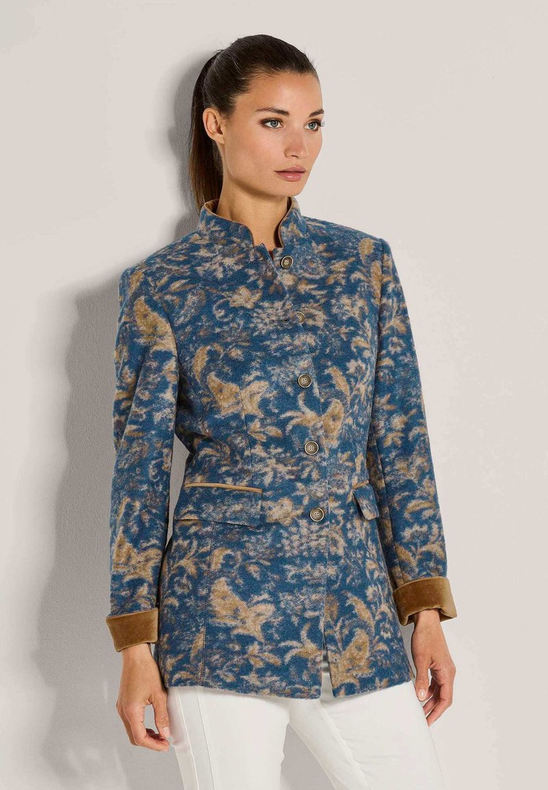 Vrouw in een blauw en tan patroon jasje met knopen en bruine manchetten, gecombineerd met een witte broek, staande tegen een beige muur.