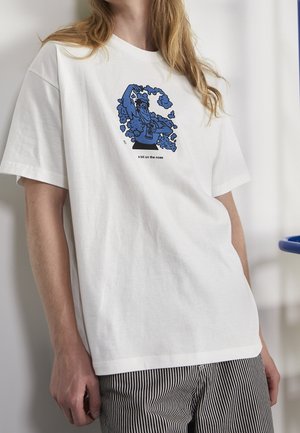 Persoon in een wit T-shirt met een blauw cartoonfiguur dat rook blaast en de tekst "een beetje voor de hand liggend", gecombineerd met zwart-wit gestreepte broek.