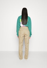 Felpa verde con stampa grafica, abbinata a pantaloni beige a vita alta. Capelli lunghi e lisci e uno sfondo minimalista.