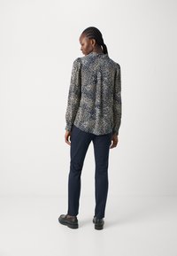 Long-sleeve blouse in navy with a cream and gold dot pattern. Features a buttoned collar and gathered cuffs, paired with navy trousers and black shoes.