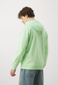 Sweatshirt verde claro com bolso canguru frontal e capuz ajustável com cordão, combinado com jeans azul. Textura de tecido suave e casual.