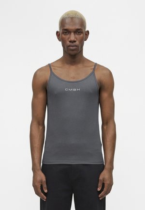 Tânăr cu păr blond scurt, purtând un maiou gri închis, mulat, cu textul "CMBH", stând pe un fundal alb simplu.