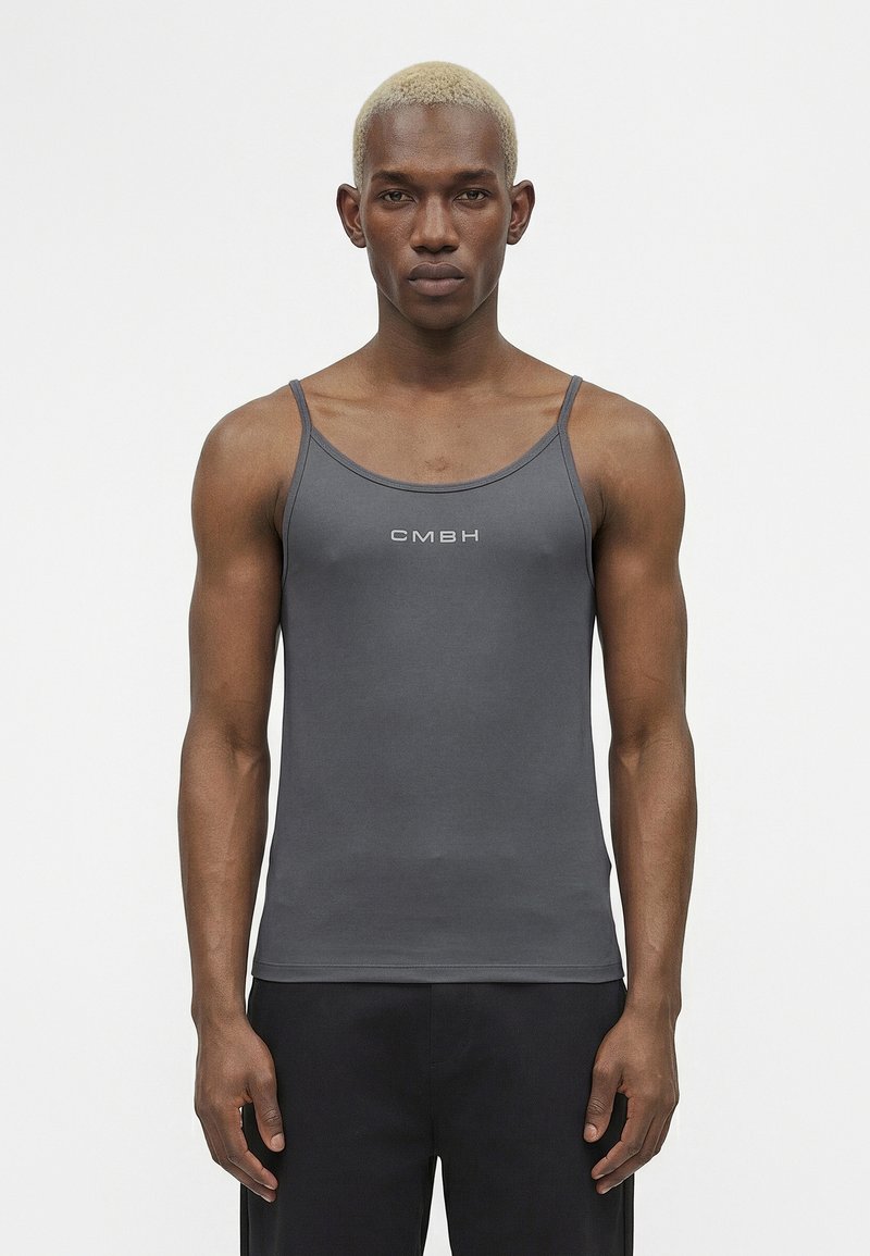 Νέος άνδρας με κοντά ξανθά μαλλιά, φοράει στενό σκούρο γκρι τοπ με την επιγραφή "CMBH" και στέκεται μπροστά σε απλό λευκό φόντο.