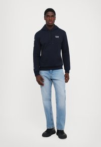 Felpa blu navy con cappuccio e cordino, tasca frontale e logo bianco sulle maniche, abbinata a jeans azzurri e scarpe da ginnastica nere.
