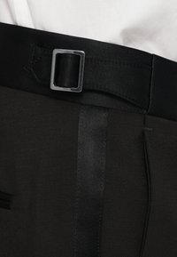 Ceinture noire réglable à boucle latérale portée sur un pantalon taillé sombre, accompagnée d'une chemise blanche.