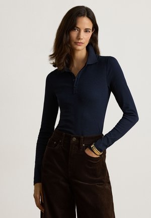 Donna con capelli scuri che indossa una camicia blu navy a maniche lunghe con colletto e pantaloni marrone scuro, in piedi con una mano in tasca su uno sfondo semplice.