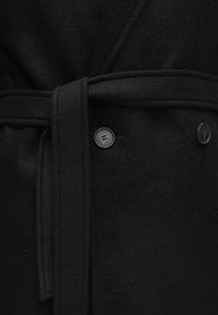 Fekete gyapjúkabát átkötős kialakítással, szövet övvel és két fekete gombbal elöl. Az anyag puha és meleg tapintású.