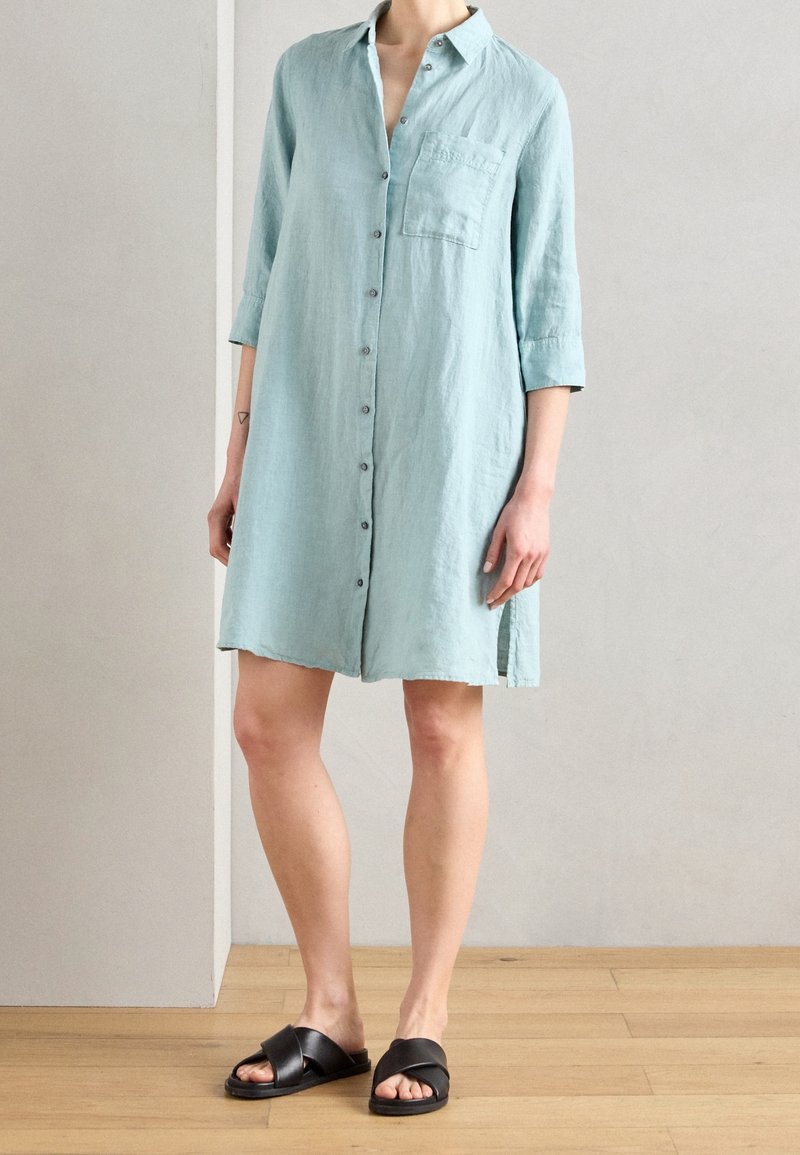 Femme portant une robe-chemise bleu clair à boutons avec des manches trois quarts et des sandales noires à brides croisées, debout sur un sol en bois.