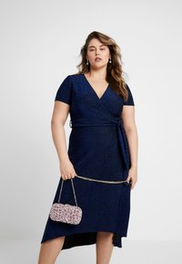 Robe cache-cœur bleu marine à manches courtes et décolleté en V, avec une texture pailletée et une taille nouée. Un petit sac à main en sequins roses vient compléter la tenue.