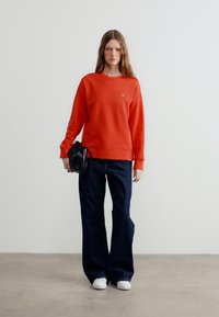 Γυναίκα όρθια με πορτοκαλί φούτερ, σκούρο τζιν με φαρδιά μπατζάκια, λευκά αθλητικά παπούτσια και κρατώντας μια μικρή μαύρη τσάντα σε απλό φόντο.