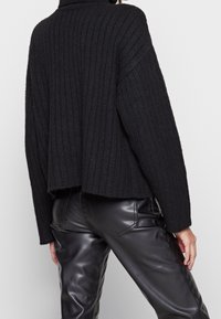 Schwarzer gerippter Strickpullover mit hohem Kragen und cropped Design, kombiniert mit glänzenden schwarzen Lederhosen. Das strukturierte Material wirkt sichtbar weich.