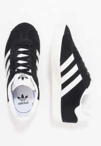 Svarta mockaskor med vita gummisulor, med tre vita ränder, rund tå och snörningsdesign. Adidas-logga på tungan.