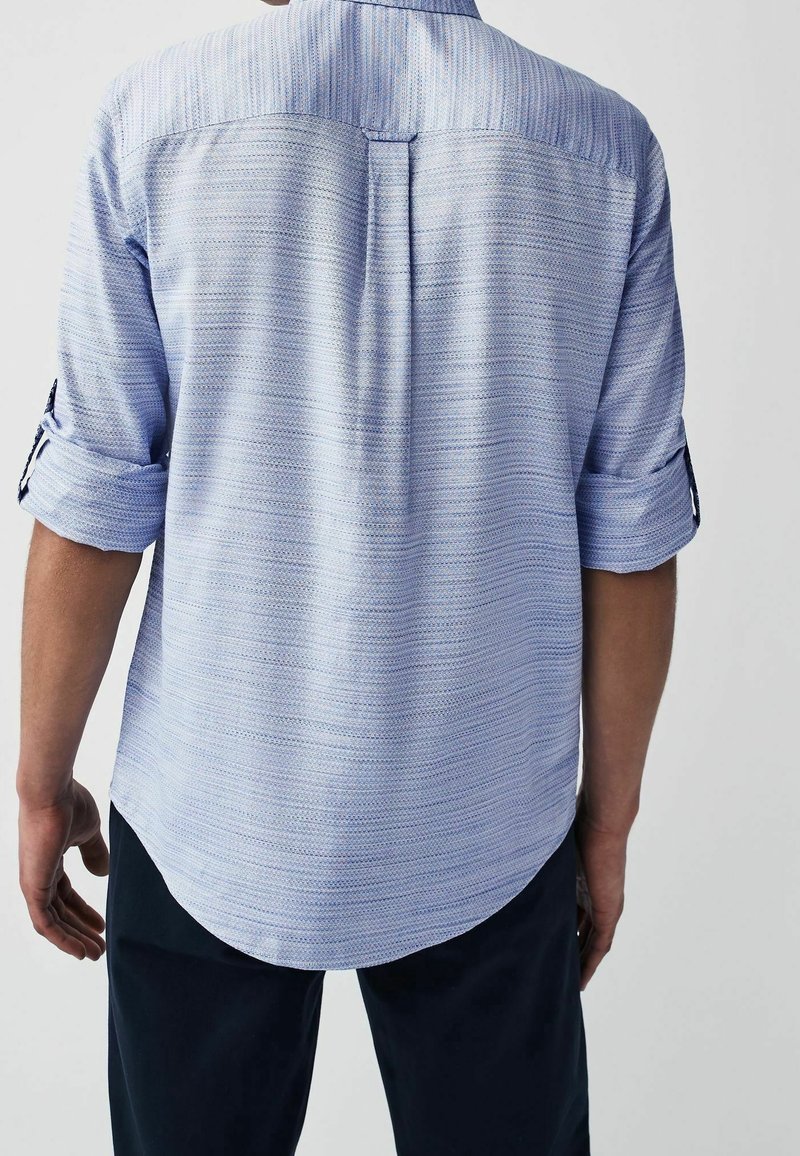Homme portant une chemise bleu clair à manches longues retroussées et un pantalon sombre, vu de dos sur un fond uni.