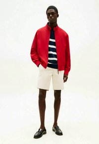 Man wearing red zip-up jacket, navy and white striped shirt, beige shorts, and black loafers, standing against plain background.