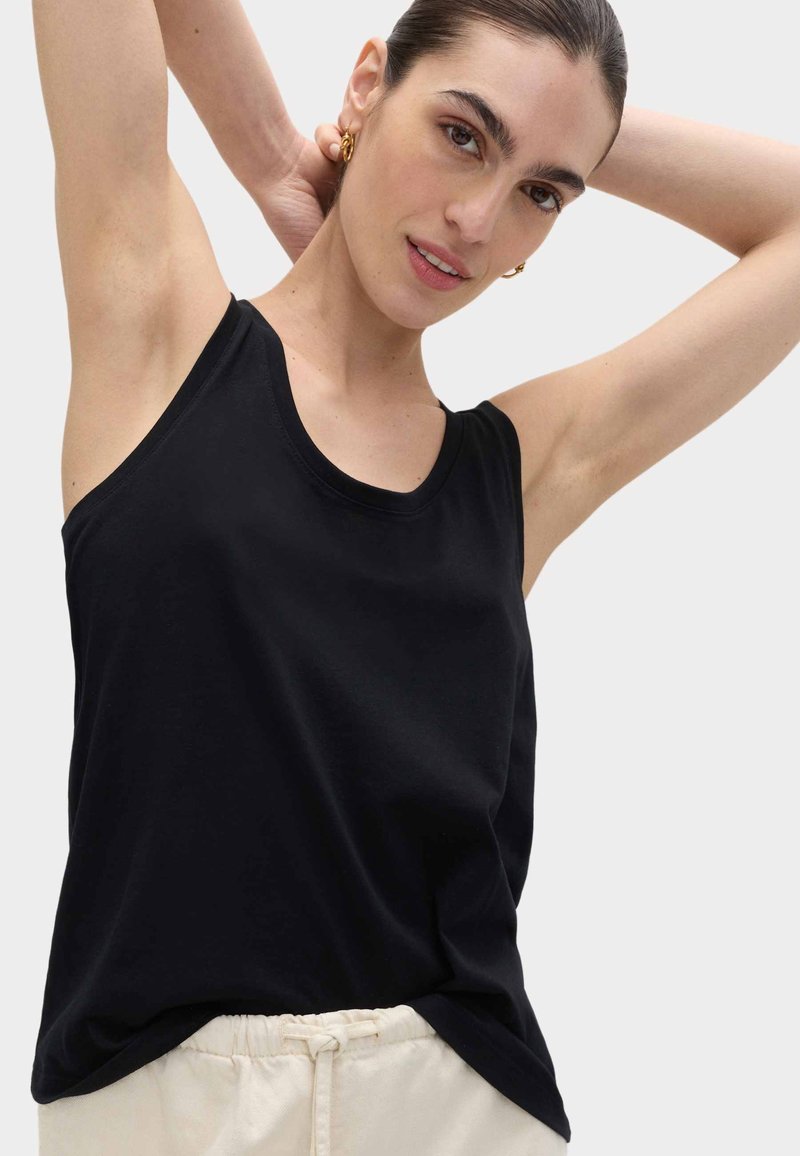 Young woman with dark hair pulled back wearing gold hoop earrings, a black sleeveless top, and light-colored pants with drawstring.