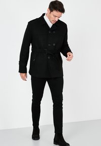 Abrigo marinero negro con botones de doble botonadura, cuello ancho y cintura con cinturón, combinado con pantalones negros ajustados y botines.