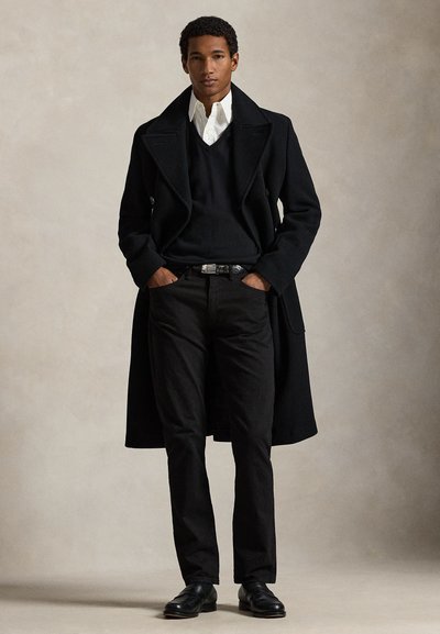 Junger Mann, der mit den Händen in den Taschen steht, trägt einen schwarzen Mantel, schwarzen Pullover, weißes Hemd, schwarze Hose und schwarze Schuhe vor einfarbigem Hintergrund.