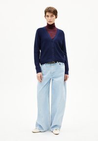 Marineblauwe cardigan met v-hals en knopen, over een bordeauxrode turtleneck gedragen. Lichtblauwe brede jeans en crèmekleurige schoenen maken de look compleet.