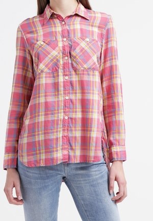 Mujer con una camisa de cuadros abotonada en rosa, naranja y amarillo con dos bolsillos en el pecho y vaqueros azul claro.
