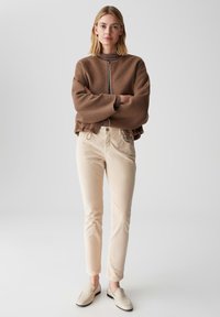 Bruine cropped jas, beige fitted broek, lichte schoenen. De jas heeft een eenvoudige ritssluiting en een zachte textuur. De broek is glad en op maat gemaakt.
