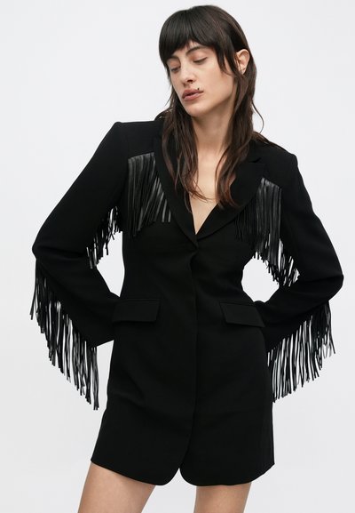 Nő fekete blézerruhában, hosszú fekete rojtozással az ujjakon és a mellrészen, kezét csípőjén tartva, egyszínű háttér előtt áll.