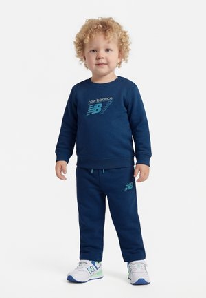 Bambino piccolo con capelli biondi ricci che indossa una felpa e pantaloni blu navy New Balance, scarpe da ginnastica bianche, in piedi su sfondo bianco.
