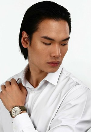 Silver chronograph watch with a textured white face and olive green strap worn on a wrist against a white dress shirt.