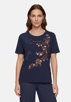 Femme portant un t-shirt bleu marine avec des motifs floraux en cuivre et le texte "Beauty of Nature" sur le devant, debout devant un fond uni.