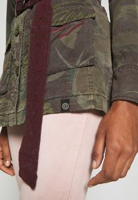 Kurtka w wzór kamuflażu w odcieniach zieleni, brązu i czerwieni. Posiada bordowy siatkowy pasek, dwie kieszenie z przodu oraz metkę z logo na dolnym brzegu.