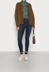 Brun fluffig kofta över en grön mönstrad polotröja, parat med mörka skinny jeans och vita sneakers. Håller en liten vinröd handväska.