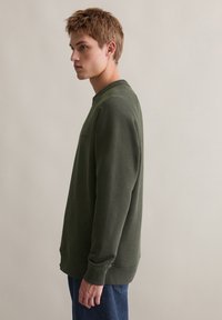 Zielona bluza z długim rękawem i okrągłym dekoltem, z ribbowanymi mankietami i dołem. Posiada delikatną teksturę i luźny krój. Nosi się ją z niebieskimi dżinsami.