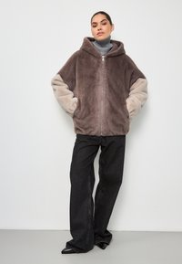 Hoodie in finta pelliccia marrone e beige, con zip frontale, vestibilità oversize e texture morbida. Abbinato a jeans a gamba larga neri.