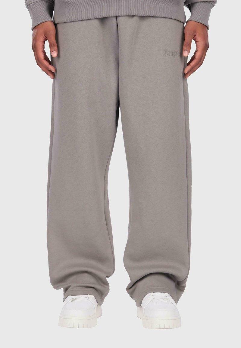 Person in lockeren grauen Sweatpants und passendem Sweatshirt, entspannt mit Händen stehend, kombiniert mit weißen Sneakers vor einem einfarbigen Hintergrund.