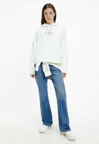 Felpa bianca con cappuccio e logo dorato, abbinata a jeans a gamba larga blu e sneakers bianche chunky. Look casual e fit rilassato.