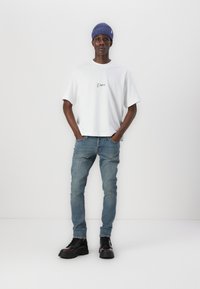 White t-shirt with black logo, light blue skinny jeans, and black chunky boots. Model wears a ribbed blue beanie. Minimalist design.