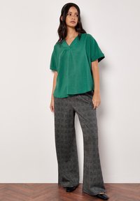 Top verde a maniche corte con scollo a V realizzato in tessuto testurizzato, abbinato a pantaloni larghi a quadri grigi e scarpe nere su un pavimento di legno.