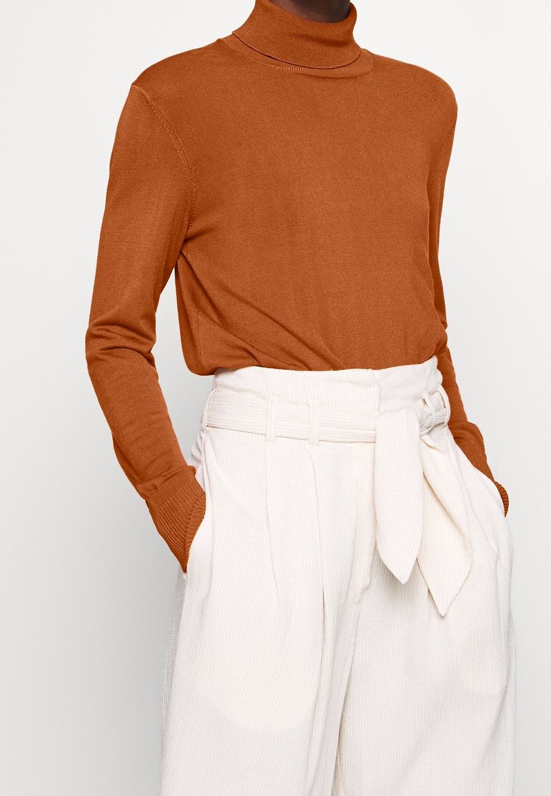 Pull à col roulé ajusté en maille orange-brun, associé à un pantalon plissé crème taille haute avec une ceinture en tissu nouée.