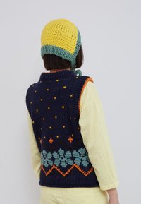 Personne portant un bonnet tricoté jaune et vert, un gilet tricoté bleu marine avec des motifs orange et bleu sarcelle, par-dessus une chemise à manches longues jaune pâle, vue de dos.