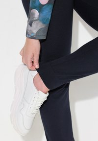 Białe skórzane sneakersy z teksturowaną podeszwą, noszone z dopasowanymi ciemnoniebieskimi spodniami. Spodnie mają prążkowany mankiet oraz kolorowy wzorzysty rękaw.