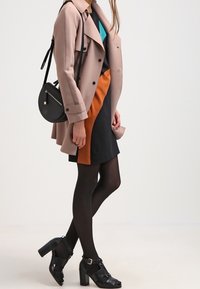 Un trench-coat beige sur une robe multicolore comportant du noir, de l'orange et du teal. Des sandales à talons noires et un sac à bandoulière noir en forme ronde.