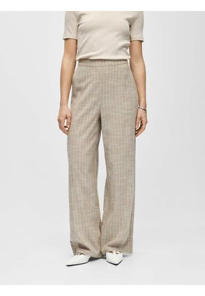 Kvinne iført beige vid bukse med smale striper, kortermet lys beige topp og hvite spisse sko mot ensfarget bakgrunn.